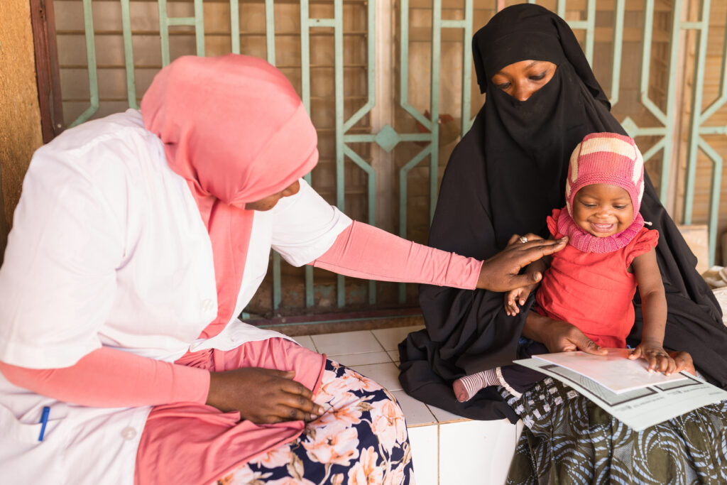 A woman wearing a burqa sits with a smiling baby on her lap. Another woman wearing a head scarf and a white medical coat examines the baby.