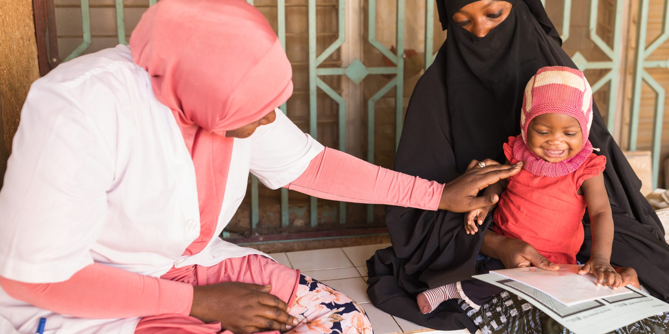 A woman wearing a burqa sits with a smiling baby on her lap. Another woman wearing a head scarf and a white medical coat examines the baby.