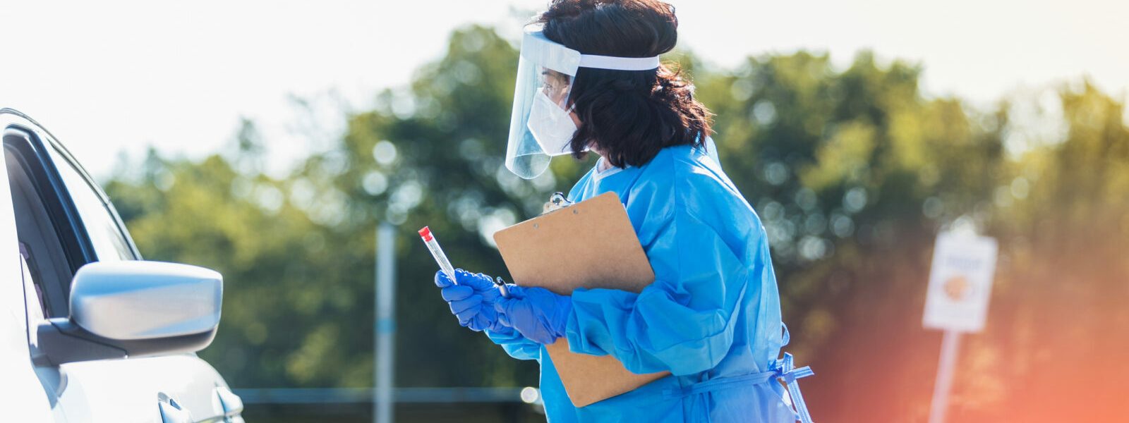 A female nurse walks up to a patient who is waiting in a car at a drive through COVID testing site. She is holding a testing kit and a clipboard. She is wearing PPE>