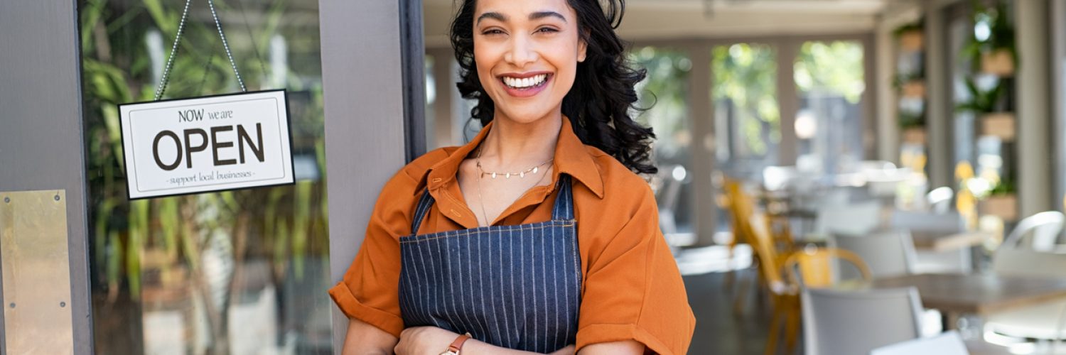 Portrait of happy waitress standing at restaurant entrance and looking at camera. Young business woman wearing apron standing with open sign at entrance gate while waiting for clients. Smiling young business owner standing at doorway of her store.