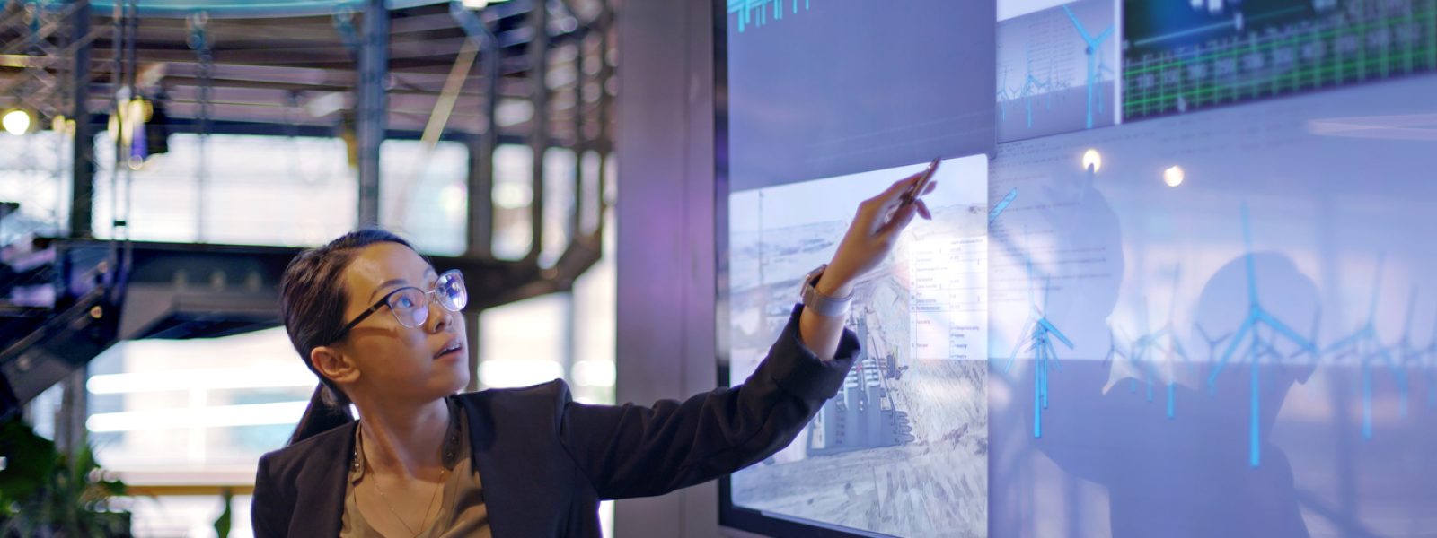 Stock photograph of a young Asian woman conducting a seminar / lecture with the aid of a large screen. The screen is displaying data & designs concerning low carbon electricity production with solar panels & wind turbines. These are juxtaposed with an image of conventional fossil fuel oil production.